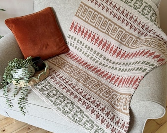 Puede incluir: Una manta estampada de color crema sobre un sillón gris. La manta presenta diseños geométricos en rojo, marrón y verde. Un cojín de terciopelo color óxido descansa sobre la silla, con una pequeña planta y una bandeja visibles.