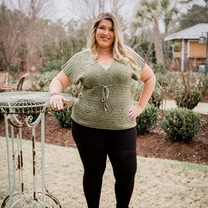 Puede incluir: Una mujer con un top de ganchillo verde con un lazo en la cintura, leggings negros y botines negros de tobillo. Est&aacute; de pie en una zona de hierba con una mesa de metal detr&aacute;s de ella.