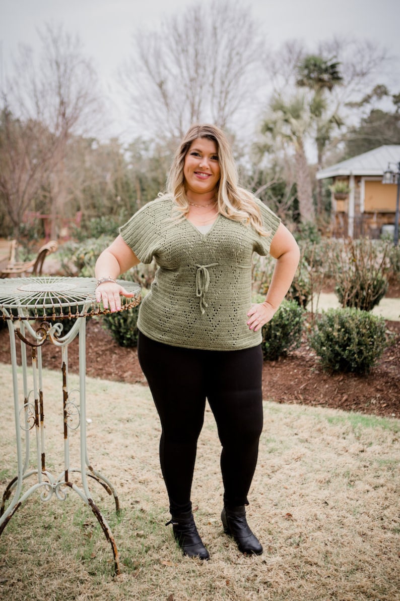 Puede incluir: Una mujer con un top de ganchillo verde con un lazo en la cintura, leggings negros y botines negros de tobillo. Est&aacute; de pie en una zona de hierba con una mesa de metal detr&aacute;s de ella.