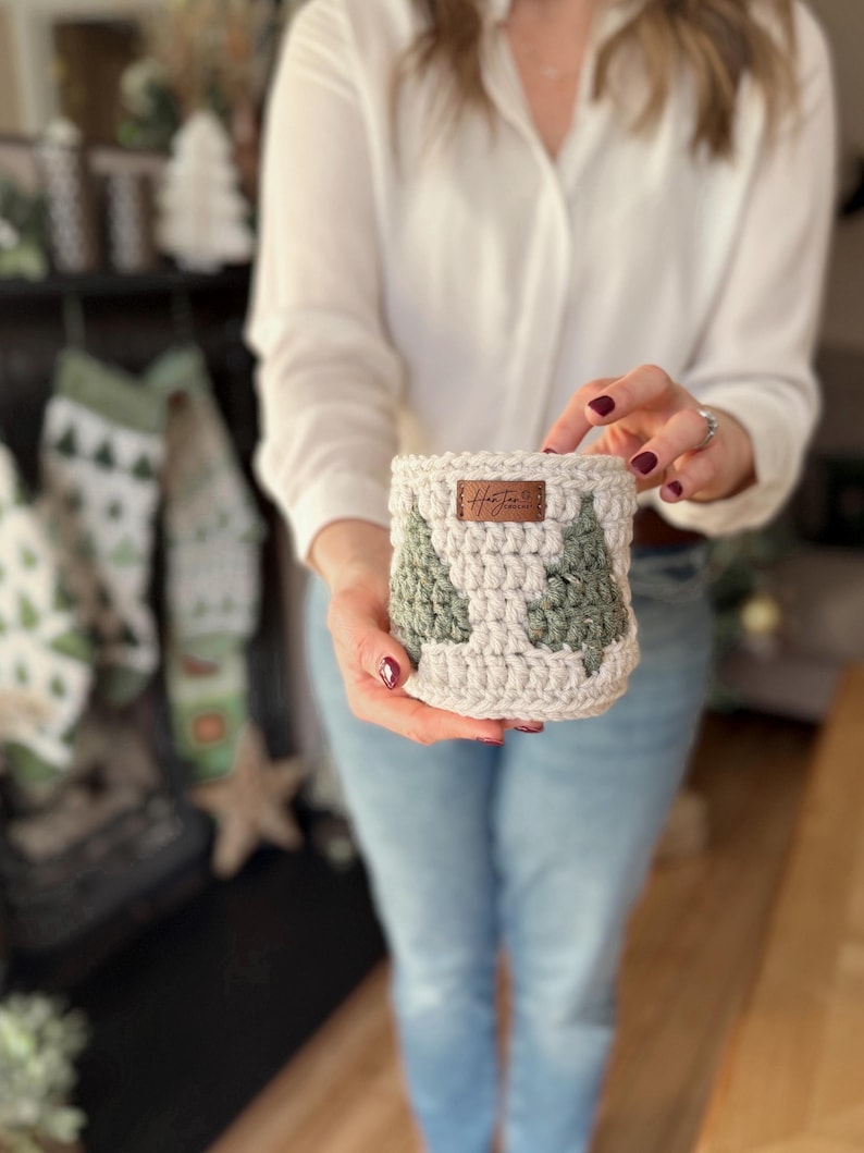 May include: A cream and green crocheted cosy with a Christmas tree design. The cosy has a small brown leather tag with the text "Hot Jar". The cosy is designed to hold a jar or cup.