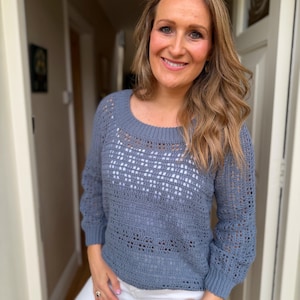 May include: A slate blue long-sleeved sweater featuring a crochet knit pattern with a round neckline and ribbed cuffs. The garment has a semi-sheer texture with alternating solid and openwork horizontal bands, paired with plain white trousers.