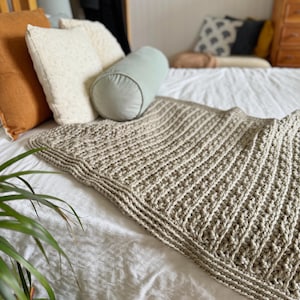 May include: A beige crocheted throw blanket with a textured pattern. The blanket is draped over a white bed with pillows.
