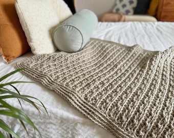 May include: A beige crocheted throw blanket with a textured pattern. The blanket is draped over a white bed with pillows.
