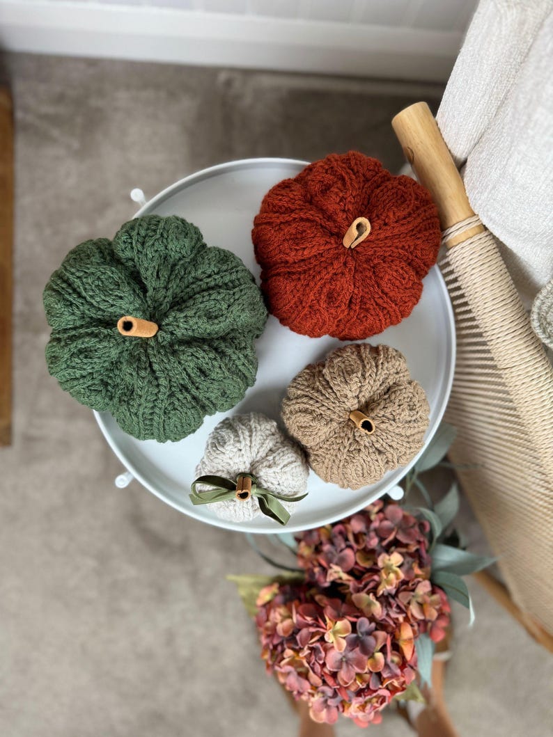 May include: Four knitted pumpkins in shades of green, orange, tan, and cream, arranged on a white tray. Each pumpkin has a wooden stem. A green ribbon adorns the smallest pumpkin. The pumpkins are a fall-themed decorative item.
