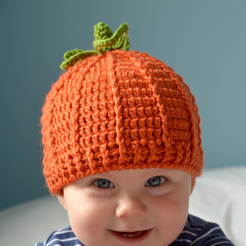 May include: A baby wearing an orange crocheted pumpkin hat with a green stem. The hat has a textured design and covers the baby's head and ears.