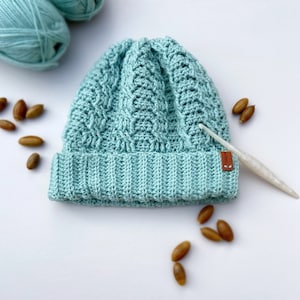 May include: A light blue crocheted beanie with a cable knit pattern. The beanie has a brown leather label with the word "Made" on it. The beanie is surrounded by acorns.