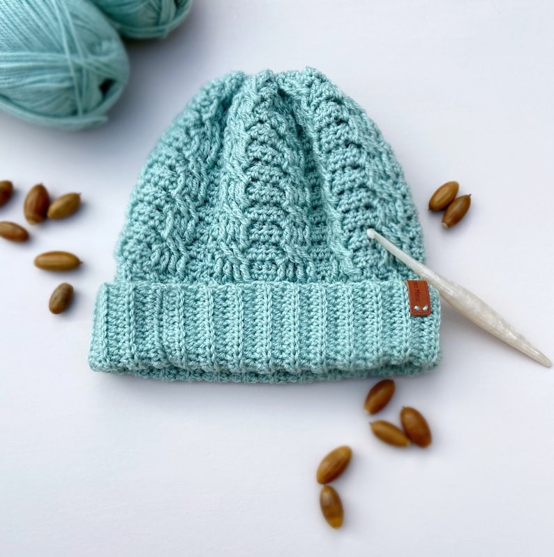 May include: A light blue crocheted beanie with a cable knit pattern. The beanie has a brown leather label with the word "Made" on it. The beanie is surrounded by acorns.