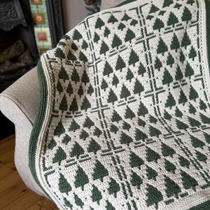 May include: A crochet blanket featuring a repeating pattern of dark green pine trees on a cream-colored background. The design is organized into a grid with thick vertical and horizontal lines, finished with a solid dark green crocheted border.