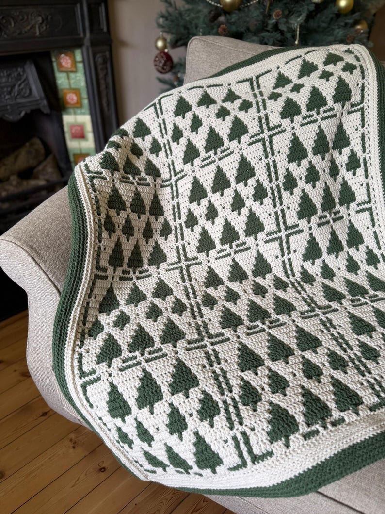 May include: A crochet blanket featuring a repeating pattern of dark green pine trees on a cream-colored background. The design is organized into a grid with thick vertical and horizontal lines, finished with a solid dark green crocheted border.