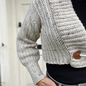 Pu&ograve; includere: Un cardigan corto con un motivo maculato beige, bianco e marrone. Il cardigan ha un collo a V e una chiusura con bottone. Il cardigan &egrave; indossato sopra una camicia nera e jeans grigi.