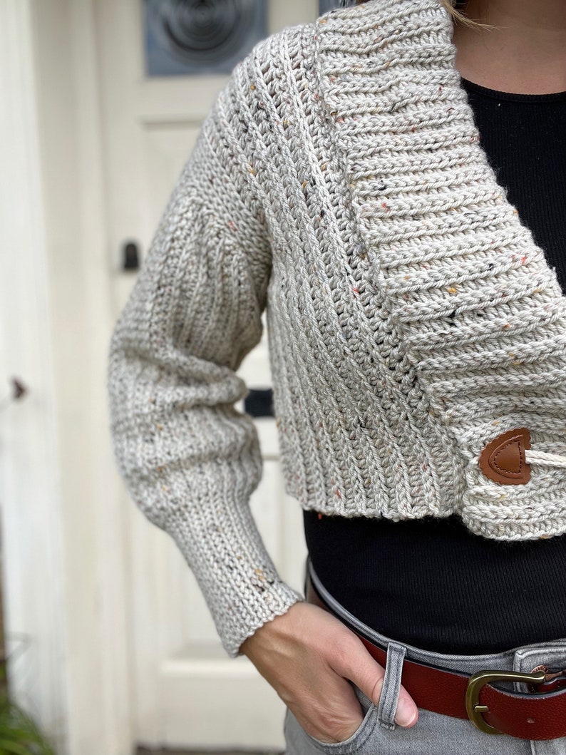 Pu&ograve; includere: Un cardigan corto con un motivo maculato beige, bianco e marrone. Il cardigan ha un collo a V e una chiusura con bottone. Il cardigan &egrave; indossato sopra una camicia nera e jeans grigi.
