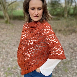 May include: Rust-colored crochet poncho with a cowl neck. The openwork design features diagonal lines and diamond shapes. The poncho is worn over a white long-sleeved shirt and blue jeans. The background is a blurred outdoor scene.