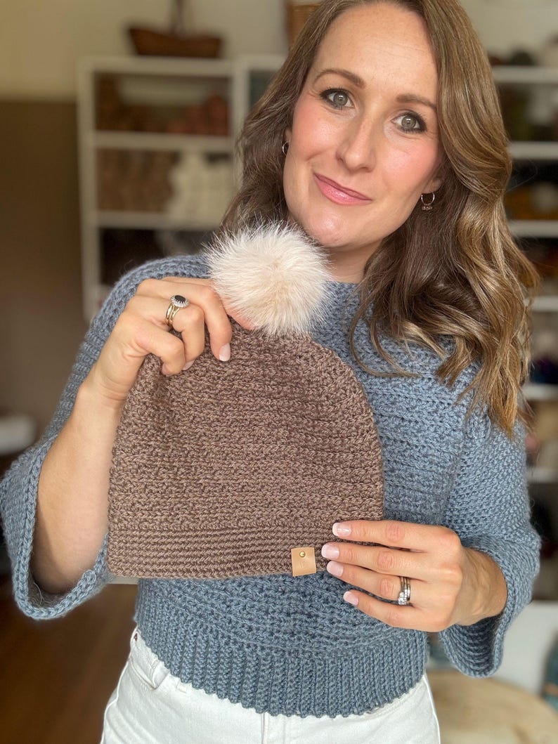 May include: A brown knit beanie with a faux fur pom-pom. The hat has a small leather tag and is held up in front of a person wearing a blue sweater. The beanie is a warm, cozy accessory.
