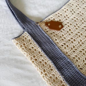 Puede incluir: Un bolso de ganchillo con un patr&oacute;n calado de color crema y bordes grises s&oacute;lidos. El bolso incluye un asa gris y un tirador de cremallera de imitaci&oacute;n cuero marr&oacute;n con herrajes de metal dorado en el borde superior del bolso.