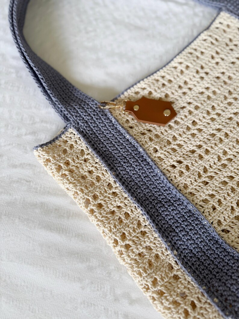 Puede incluir: Un bolso de ganchillo con un patr&oacute;n calado de color crema y bordes grises s&oacute;lidos. El bolso incluye un asa gris y un tirador de cremallera de imitaci&oacute;n cuero marr&oacute;n con herrajes de metal dorado en el borde superior del bolso.