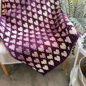Op de afbeelding: Een handgemaakte gehaakte deken met een hartpatroon in tinten paars, roze en wit. De deken is over een stoel gedrapeerd, wat de ingewikkelde steek en kleurvariaties laat zien. Een gezellig woondecoratie-item.