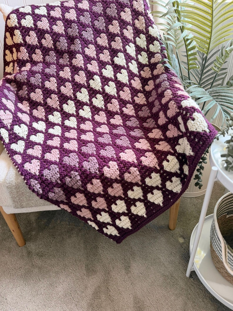 Op de afbeelding: Een handgemaakte gehaakte deken met een hartpatroon in tinten paars, roze en wit. De deken is over een stoel gedrapeerd, wat de ingewikkelde steek en kleurvariaties laat zien. Een gezellig woondecoratie-item.