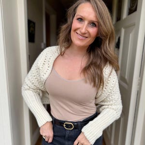 May include: Cream-colored crochet shrug with long sleeves. The shrug is worn over a beige tank top and paired with black jeans and a black belt with a gold buckle. The outfit is completed with rings.
