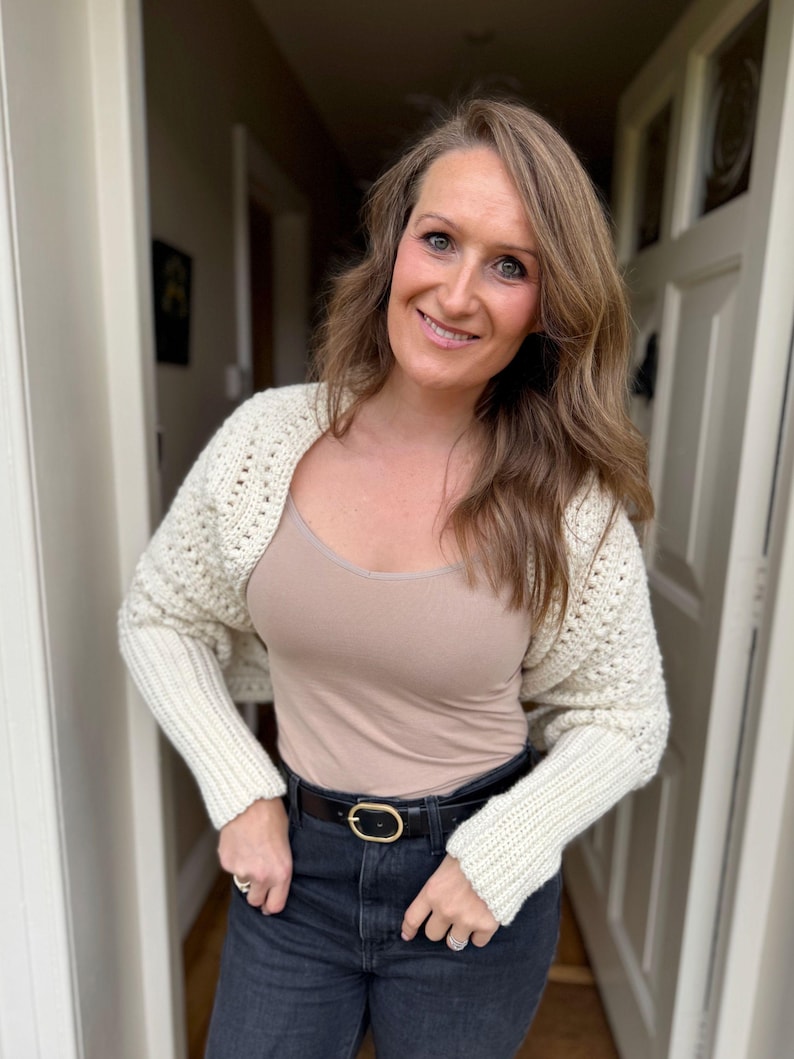 May include: Cream-colored crochet shrug with long sleeves. The shrug is worn over a beige tank top and paired with black jeans and a black belt with a gold buckle. The outfit is completed with rings.