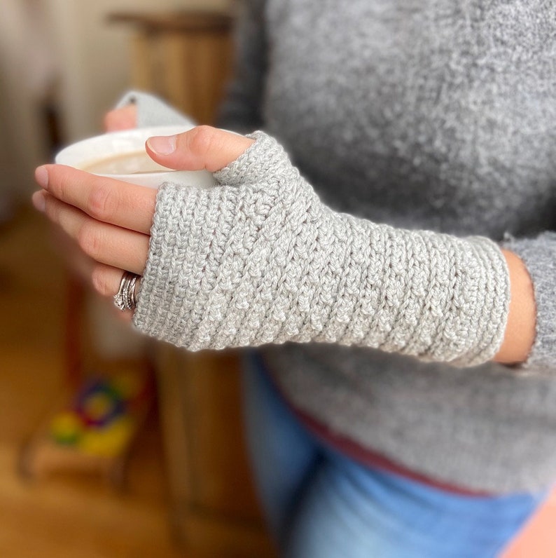 May include: Gray crocheted fingerless gloves with a ribbed texture. The gloves are worn on a person's hand, and the person is holding a white mug.