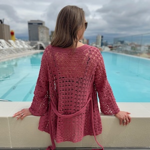 May include: A dusty rose crochet cardigan with long sleeves and a matching belt. The openwork design of the sweater is visible. The person is wearing sunglasses and is standing near a pool.