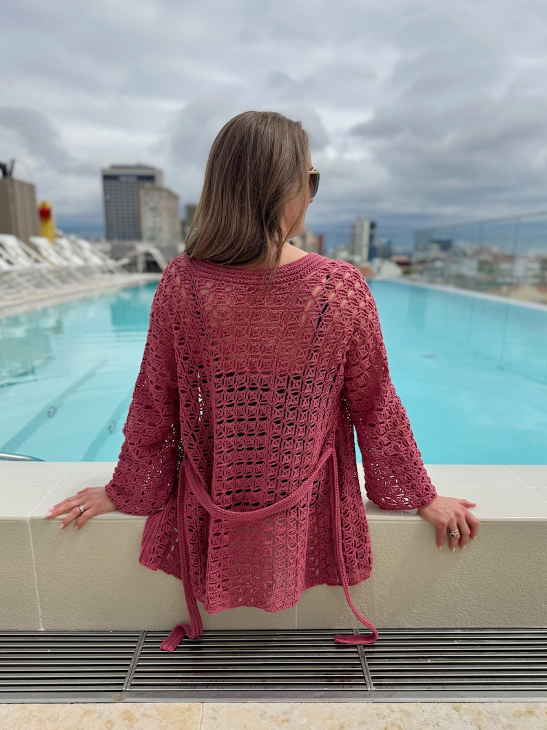 May include: A dusty rose crochet cardigan with long sleeves and a matching belt. The openwork design of the sweater is visible. The person is wearing sunglasses and is standing near a pool.