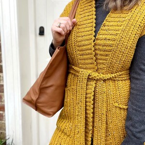 Pu&ograve; includere: Un lungo gilet di uncinetto giallo senape con scollo a V, tasche e cintura da annodare.