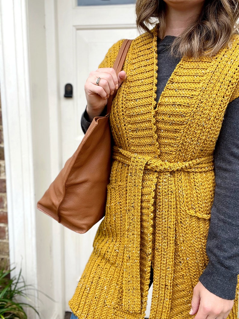 Pu&ograve; includere: Un lungo gilet di uncinetto giallo senape con scollo a V, tasche e cintura da annodare.