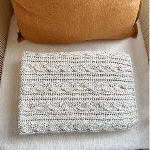 Peut inclure: Une couverture crochet&eacute;e pli&eacute;e, de couleur gris clair, avec un motif en tricot torsad&eacute;. La couverture est rectangulaire et repose sur une surface de couleur claire. Un coussin orange est visible en arri&egrave;re-plan, pos&eacute; sur une chaise.