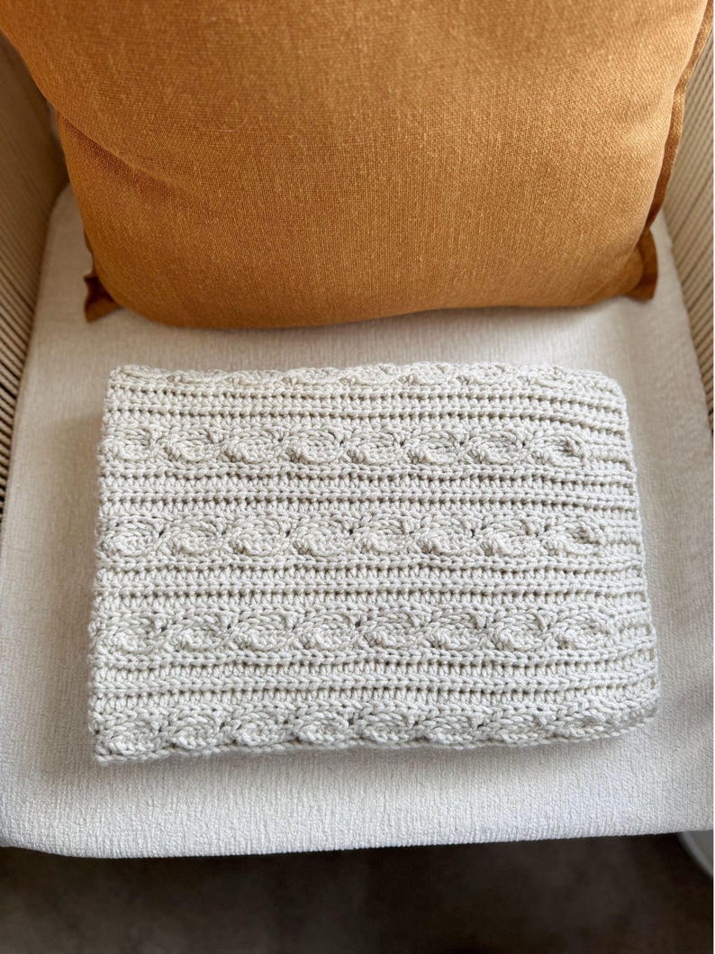 Peut inclure: Une couverture crochet&eacute;e pli&eacute;e, de couleur gris clair, avec un motif en tricot torsad&eacute;. La couverture est rectangulaire et repose sur une surface de couleur claire. Un coussin orange est visible en arri&egrave;re-plan, pos&eacute; sur une chaise.