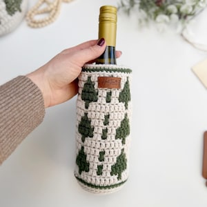 May include: A wine bottle in a cream-colored, crocheted cozy with a green tree pattern. The cozy has a brown leather tag with text. The bottle has a gold-colored top.