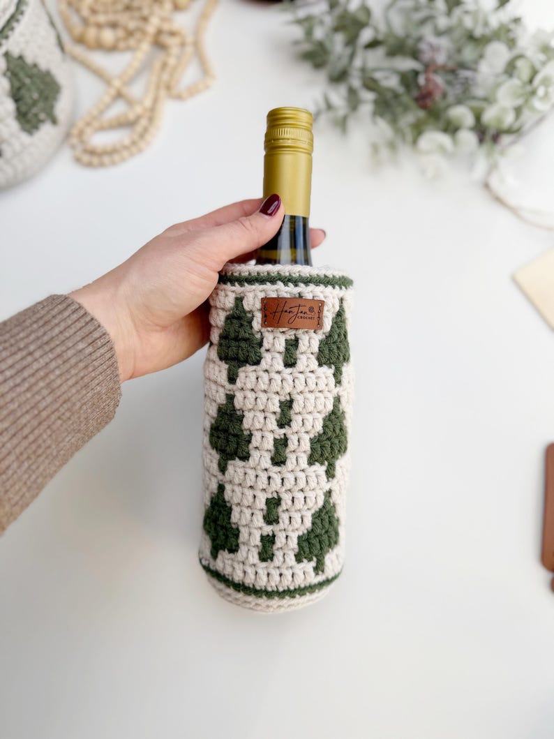 May include: A wine bottle in a cream-colored, crocheted cozy with a green tree pattern. The cozy has a brown leather tag with text. The bottle has a gold-colored top.