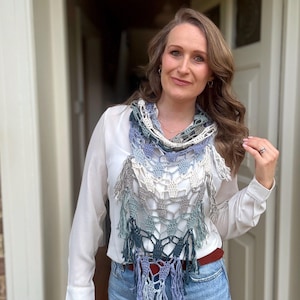 May include: A woman wearing a white shirt and blue jeans with a crocheted scarf in shades of blue, gray, and white with fringe.