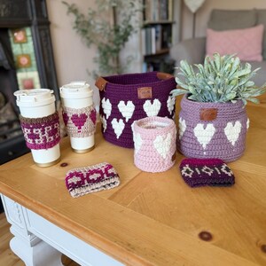 Op de afbeelding: Een verzameling handgemaakte gehaakte items, waaronder koffiebekerhoezen, manden en een plantenpotcover. De items zijn in tinten paars, roze en wit, met een hartpatroon. Ze worden getoond op een houten tafel.