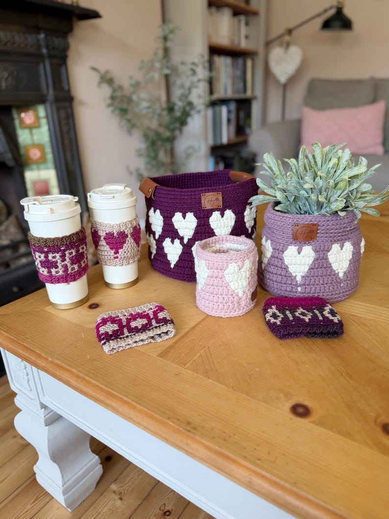 Op de afbeelding: Een verzameling handgemaakte gehaakte items, waaronder koffiebekerhoezen, manden en een plantenpotcover. De items zijn in tinten paars, roze en wit, met een hartpatroon. Ze worden getoond op een houten tafel.