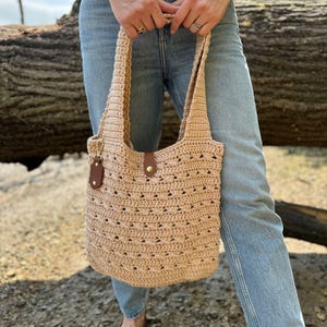 May include: A beige crochet tote bag with a leather accent and a gold-tone clasp. The bag has a textured pattern and sturdy handles. The bag is held by a person wearing blue jeans and brown shoes.