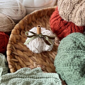 May include: A selection of knitted pumpkins in shades of cream, orange, and green, displayed in a woven basket. The image includes balls of yarn and a white pumpkin-shaped container. A small pumpkin features a cinnamon stick stem and a green ribbon bow.