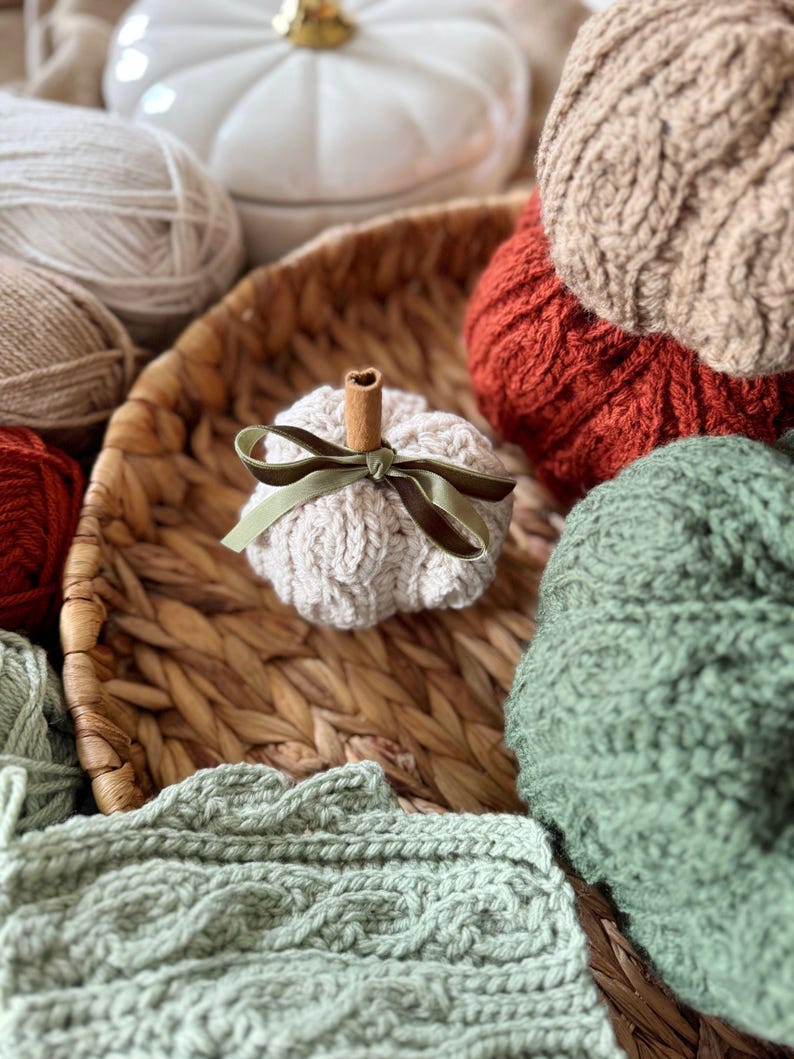 May include: A selection of knitted pumpkins in shades of cream, orange, and green, displayed in a woven basket. The image includes balls of yarn and a white pumpkin-shaped container. A small pumpkin features a cinnamon stick stem and a green ribbon bow.