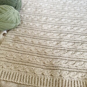 Peut inclure: Une couverture beige tricot&eacute;e avec un motif en torsade. La couverture a une bordure c&ocirc;tel&eacute;e et est faite de fil doux. Plusieurs pelotes de fil de couleurs vertes et beiges sont dans le coin sup&eacute;rieur gauche.