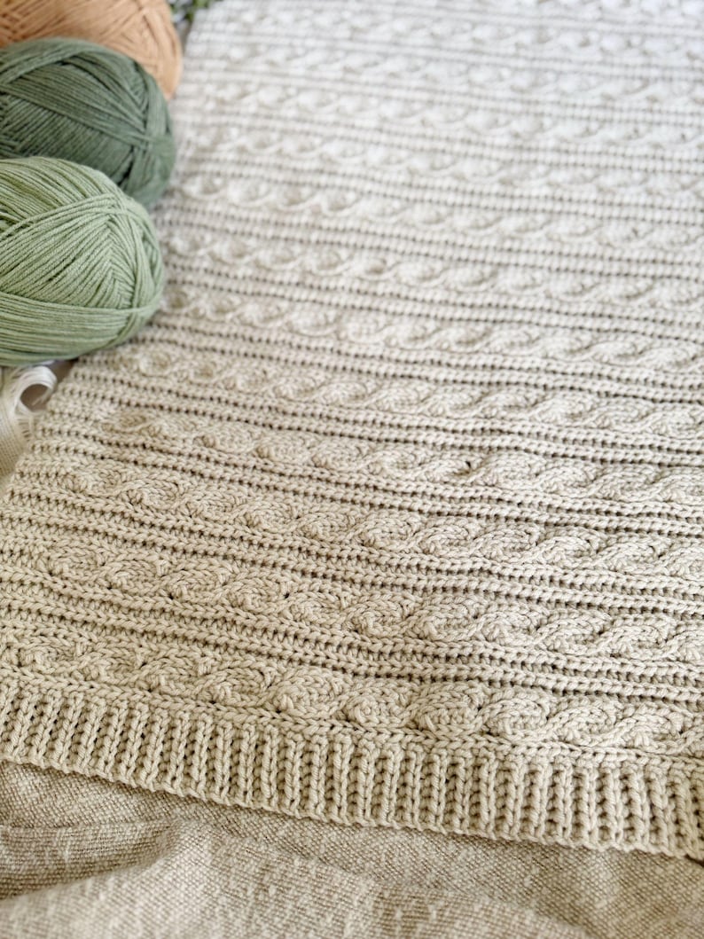 Peut inclure: Une couverture beige tricot&eacute;e avec un motif en torsade. La couverture a une bordure c&ocirc;tel&eacute;e et est faite de fil doux. Plusieurs pelotes de fil de couleurs vertes et beiges sont dans le coin sup&eacute;rieur gauche.