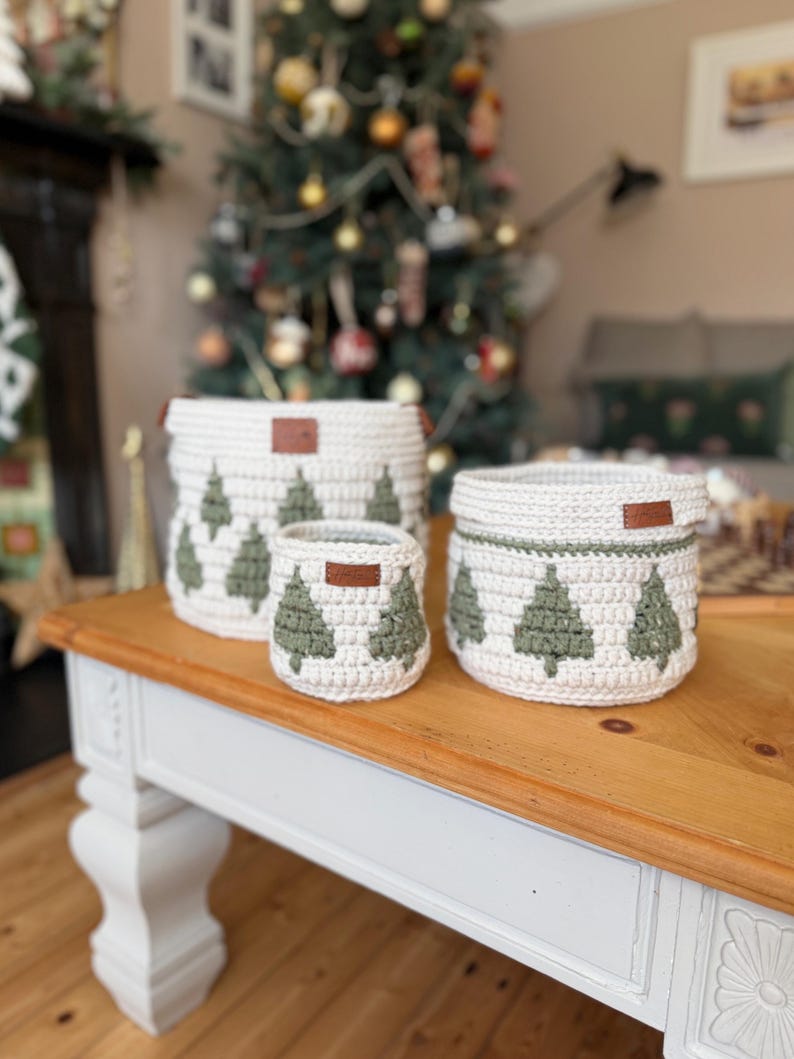 May include: Three white crochet baskets with green Christmas tree patterns and brown leather tags. The baskets are different sizes and are on a wooden coffee table. A decorated Christmas tree is in the background.