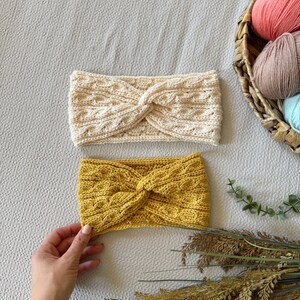 May include: Two knitted headbands in cream and mustard yellow. The headbands have a twisted knot design and a textured cable knit pattern. A basket of yarn in various pastel colors is in the background.