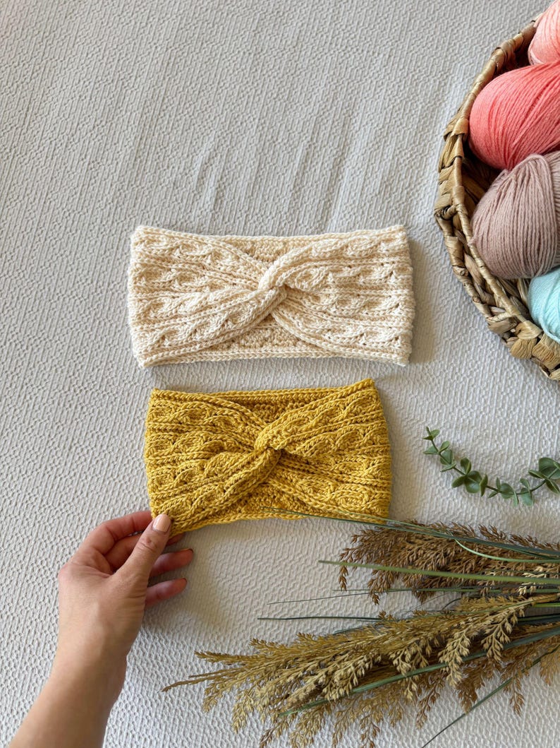 May include: Two knitted headbands in cream and mustard yellow. The headbands have a twisted knot design and a textured cable knit pattern. A basket of yarn in various pastel colors is in the background.