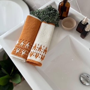 May include: Two handmade crochet hand towels. One is orange with a white and orange geometric pattern, and the other is white with an orange and white geometric pattern. The towels are displayed on a white sink.