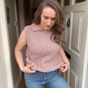 May include: A sleeveless, dusky pink knitted top with a collared neckline. The top is worn with light blue denim jeans. The knitted texture is visible, and the overall style is casual and comfortable.