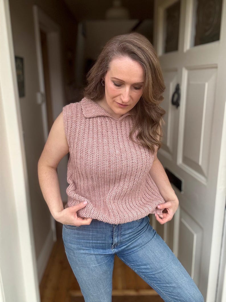 May include: A sleeveless, dusky pink knitted top with a collared neckline. The top is worn with light blue denim jeans. The knitted texture is visible, and the overall style is casual and comfortable.