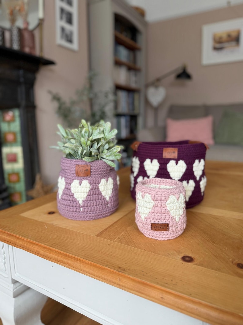 Op de afbeelding: Drie gehaakte manden met hartpatronen in verschillende maten en tinten paars en roze staan op een houten tafel. De manden hebben witte hartontwerpen en kleine bruine leren labels. E&eacute;n mand bevat een groene plant.