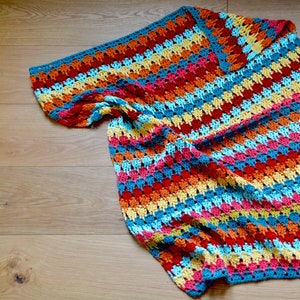 Könnte beinhalten: Eine bunte gehäkelte Wolldecke mit Streifen in Rot, Orange, Gelb, Blau und Rosa.