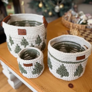 May include: A set of three crocheted baskets in different sizes. The baskets are off-white with a green tree pattern. Each basket has a brown leather tag and handles. The baskets are on a wooden surface.