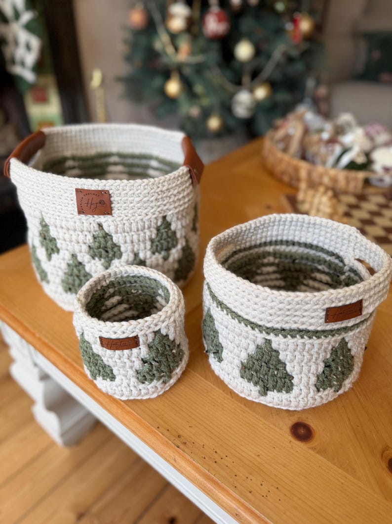 May include: A set of three crocheted baskets in different sizes. The baskets are off-white with a green tree pattern. Each basket has a brown leather tag and handles. The baskets are on a wooden surface.